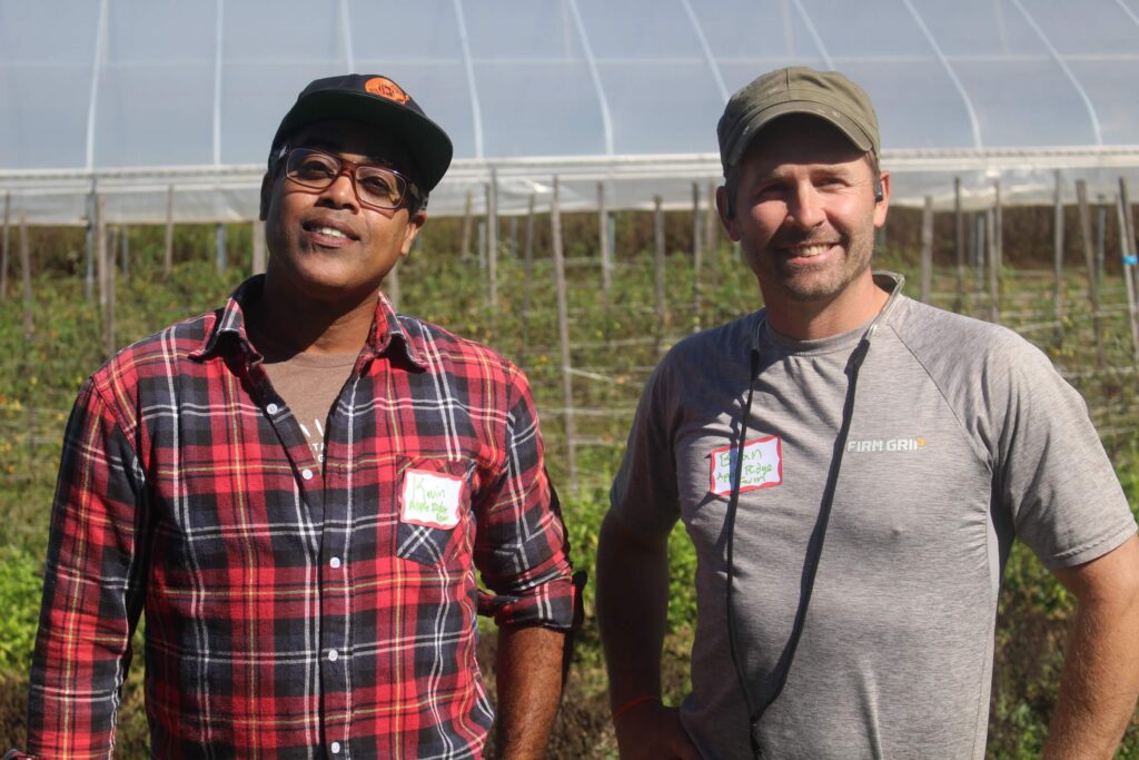 Apprentice Kevin Jackasal in a hat and red flannel and mentor farmer Brian Bruno in a hat and gray t-shirt standing next to each other in front of a hoop house and smiling at the camera