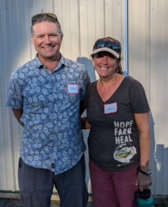 Farmers John Good in a blue button-down shirt and Aimee Good in a hat and gray t-shirt with the words "Hope, farm, heal" standing next to each other smiling at the camera