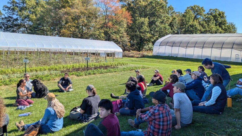 Farmers John and Aimee Good, and a black dog on a field in front of a hoop house facing a group of apprentices sitting on grass with their backs turned away from the camera