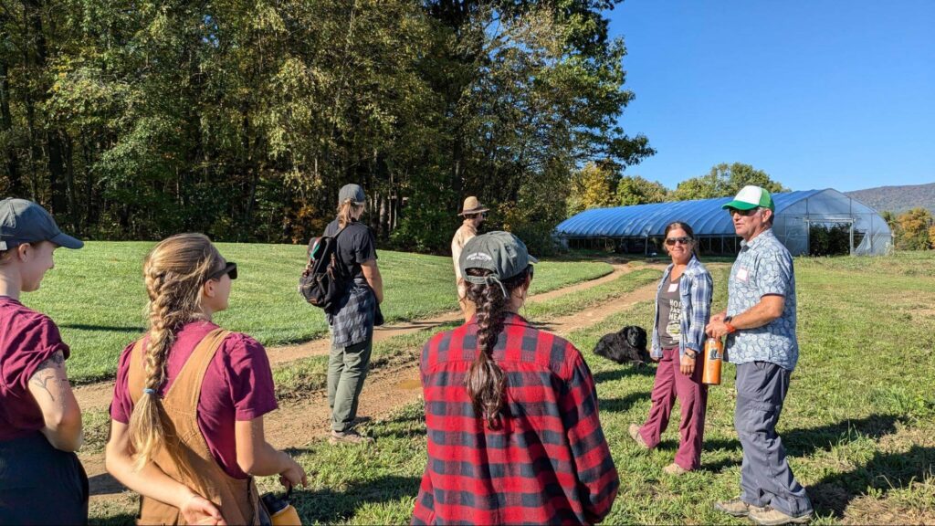 Two farmers facing a group of five apprentices with their backs turned away from the camera on a field with trees, a high tunnel, and a black dog in the background